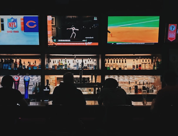A bar counter with several people sitting in front of it is shown. Above the counter, there are multiple television screens displaying sports broadcasts, including football and baseball. Behind the counter, various bottles of alcohol are neatly arranged.