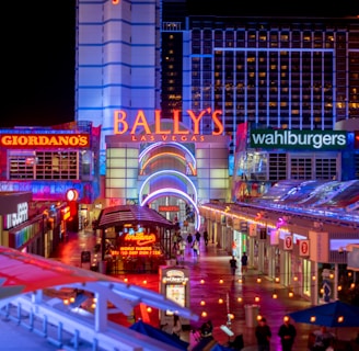 A brightly lit urban scene featuring the entrance to Bally's Las Vegas, surrounded by various illuminated shop signs including Giordano's, Wahlburgers, and Redneck Riviera. The street is bustling with people and rich with neon lights and colorful signage, creating a lively nighttime atmosphere.