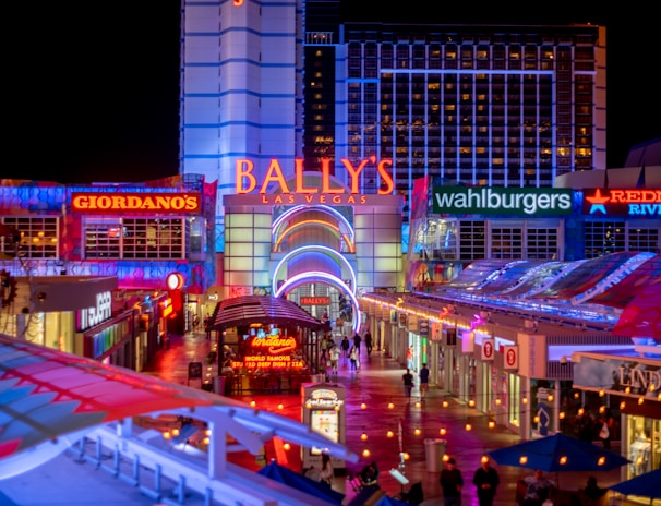 A brightly lit urban scene featuring the entrance to Bally's Las Vegas, surrounded by various illuminated shop signs including Giordano's, Wahlburgers, and Redneck Riviera. The street is bustling with people and rich with neon lights and colorful signage, creating a lively nighttime atmosphere.