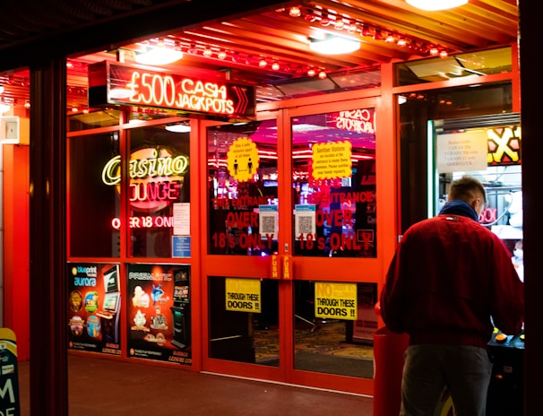 A brightly lit entrance to a casino with neon signs, including one advertising a £500 jackpot. The doors have clear signage indicating entry is restricted to those over 18. A person in a red jacket stands to the right, possibly interacting with a machine or display.