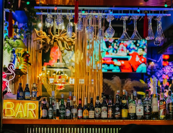 An illuminated bar counter features a variety of liquor bottles, with colorful neon lights adding vibrancy. Glasses hang upside down above the bar, and a large television screen in the background displays a sports event.