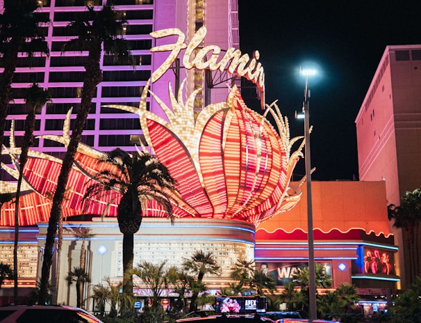 A brightly lit casino with neon lights in vibrant red, pink, and blue, featuring a large flamingo signage. Palm trees are visible in the foreground with vehicles passing by on the street at night.